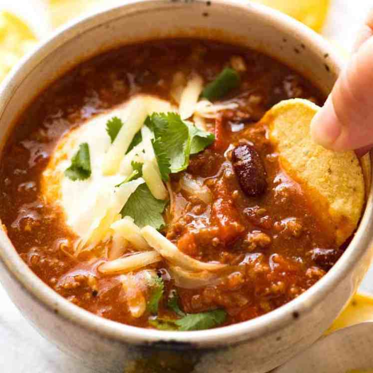 Corn chip being dipped into a bowl of Chili topped with sour cream, grated cheese and cilantro